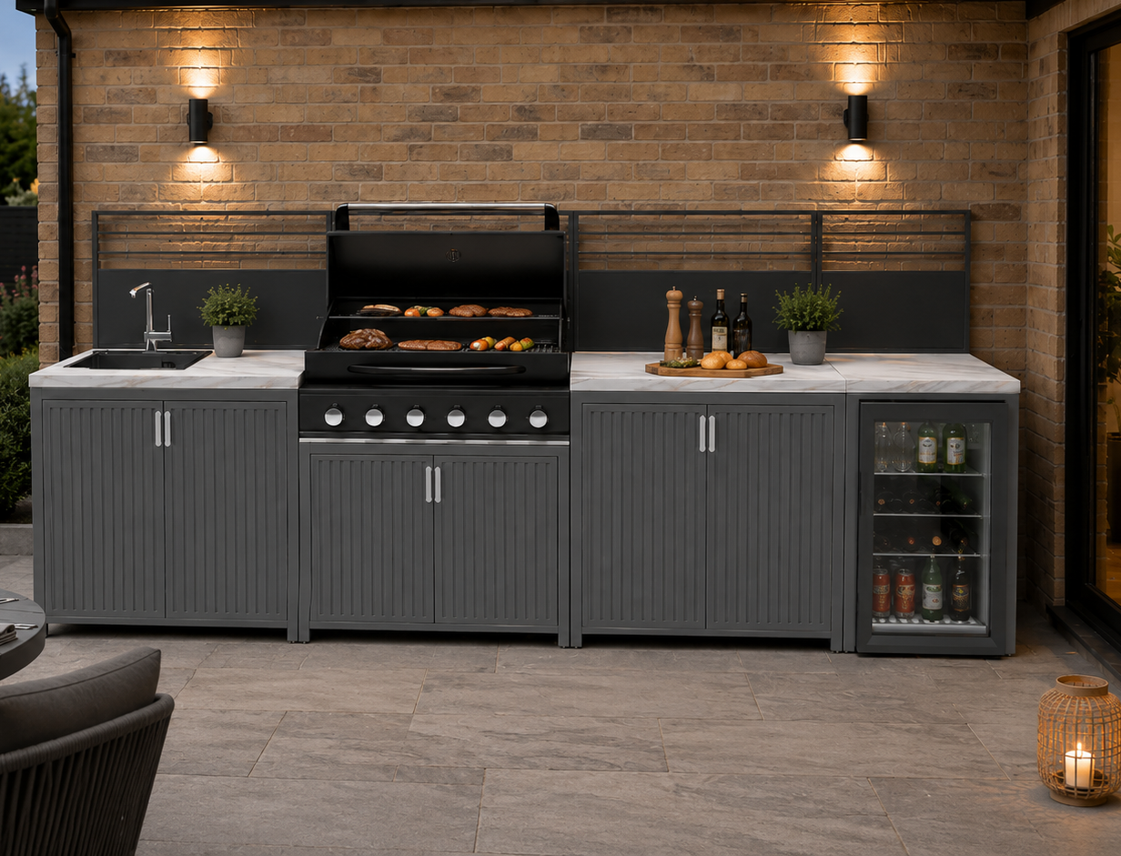 A modern outdoor kitchen island set on a paved patio, featuring gray ribbed cabinets, a black gas grill with food cooking, a sink, a countertop with bottles and accessories, and a glass-door beverage refrigerator. A dining table with candles is visible in the foreground, and the backdrop is a brick wall with wall lights.